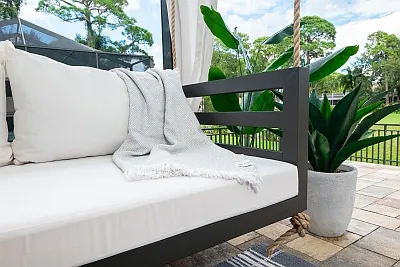 Shirley swing bed in bronze with white cushions and a gray knit throw beside a potted snake plant